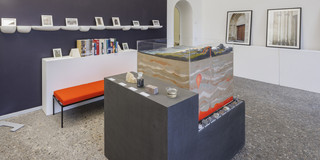 Zu sehen ist eine Natursteinausstellung. Im Zentrum des Bildes befindet sich eine Art "Ausstellungsinsel" mit verschiedenen Steinen und einer durchsichtigen Box, welches als Anschauungsmaterial dient. Links hinter der Insel an einer dunklen Wand befindet sich eine orangene Bank vor einer Art weißem Sideboard, auf dem sich ebenfalls Ausstellungsstücke befinden. Darüber hängt eine weiße Regalreihe an der Wand mit weiteren Anschauungsstücken.