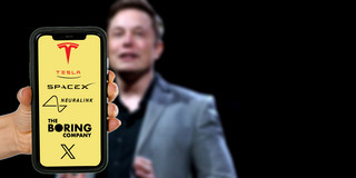 Im Hintergrund ein leicht verschwommenes Foto von Elon Musk auf einer Bühne. Im Vordergrund hält eine Hand ein Smartphone ins Bild, auf dem Display sind die Logos von Tesla, SpaceX, Neuralink, The Boring Company und X auf gelbem Hintergrund zu erkennen.