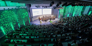 Das Bild zeigt das Audimax der TU Dortmund bei der Akademischen Jahresfeier 2024. Das Foto wurde von oben aufgenommen und zeigt das Publikum von hinten. Im Zentrum, weiter oben im Bild ist das Orchester der TU Dortmund zu sehen, darüber die Präsentation. Die seitlichen Wände des Audimax sind grün angestrahlt und werden zusätzlich auf beiden Seiten mit zwei Kreisen, in denen das TU-Logo sowie die Zahl 24 angezeigt wird, angestrahlt.