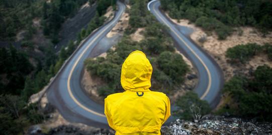 Person in einer gelben Jacke sitzt auf einen Felsen und blickt auf eine Straßen Kurve.