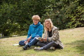 Ein blonder Student mit blauer Jacke und eine blonde Studentin mit brauner Jacke sitzen auf einer Wiese